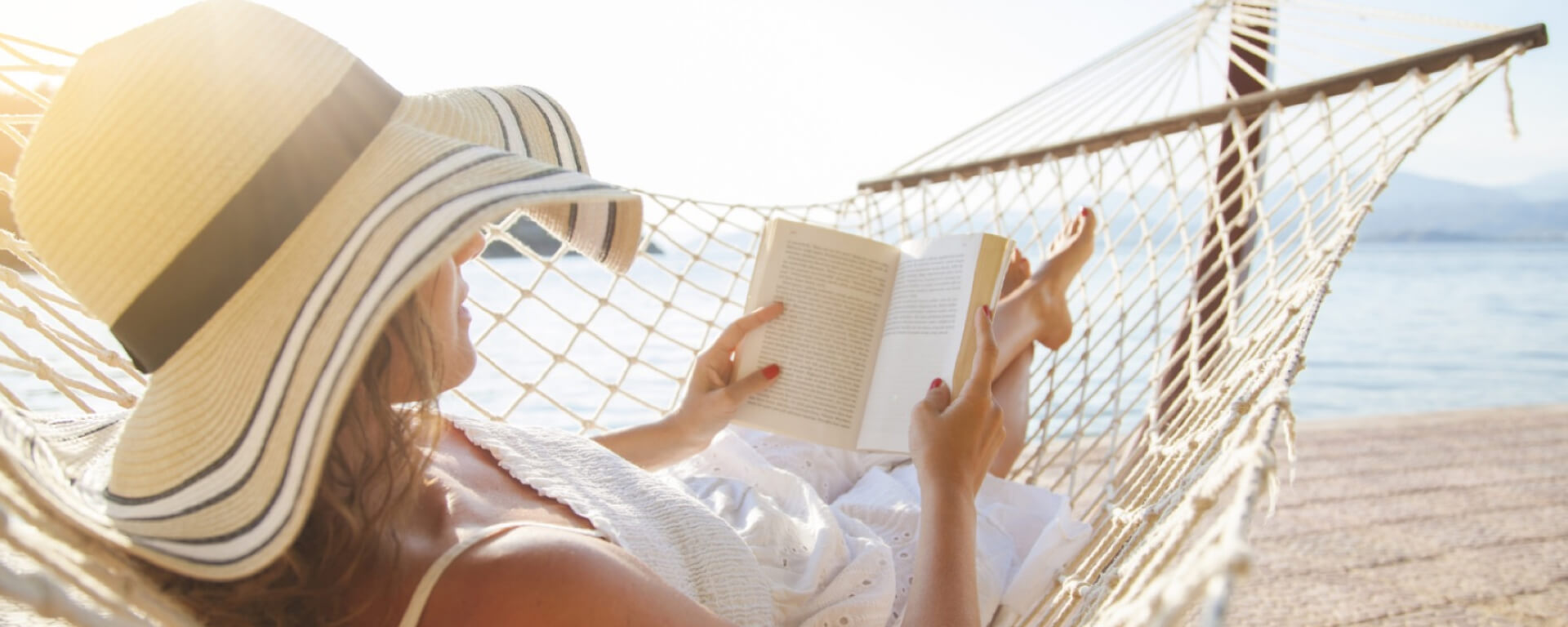Woman relaxing in a hammock on the beach, reading a book under a wide-brimmed sun hat.