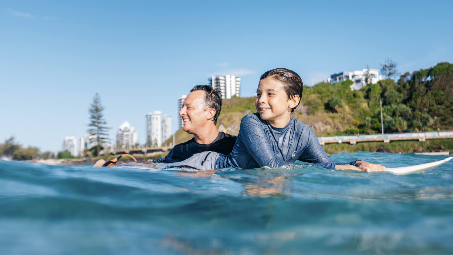 Father and son enjoying a surf session in the ocean near Coolangatta, with coastal buildings in the background.