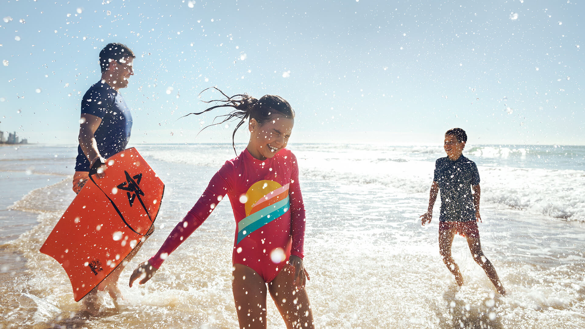 Children and an adult enjoying the surf at the beach, with bright sunlight and splashing water creating a fun, energetic scene.