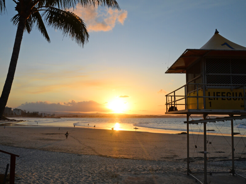 Golden sunset at Rainbow Bay Beach with a lifeguard tower and beachgoers along the water.