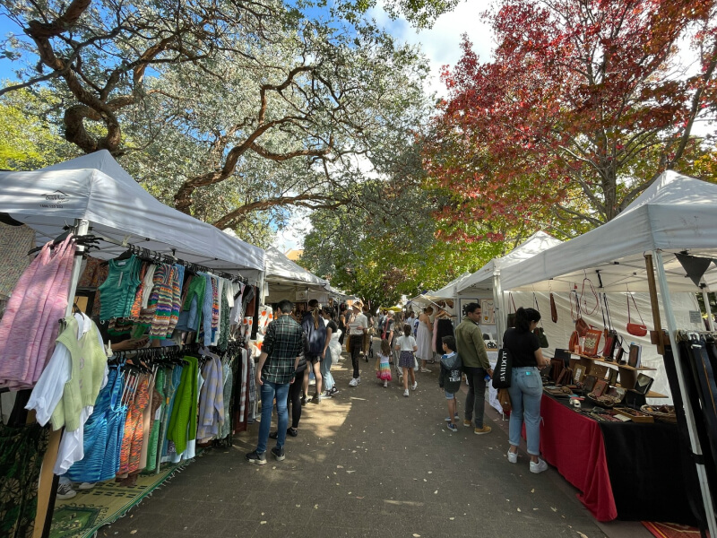 People browsing stalls at the Tweed Heads Markets, surrounded by large leafy trees and white tents displaying local crafts, clothing, and produce.