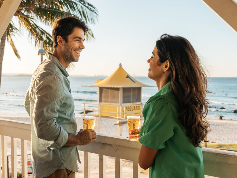 Couple enjoying drinks on a beachfront balcony at sunset, overlooking the ocean and a yellow lifeguard tower.