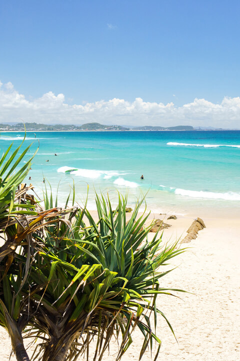 Beach scene at Coolangatta with turquoise waves, surfers in the water, and a couple relaxing under a red and blue umbrella on the sand.