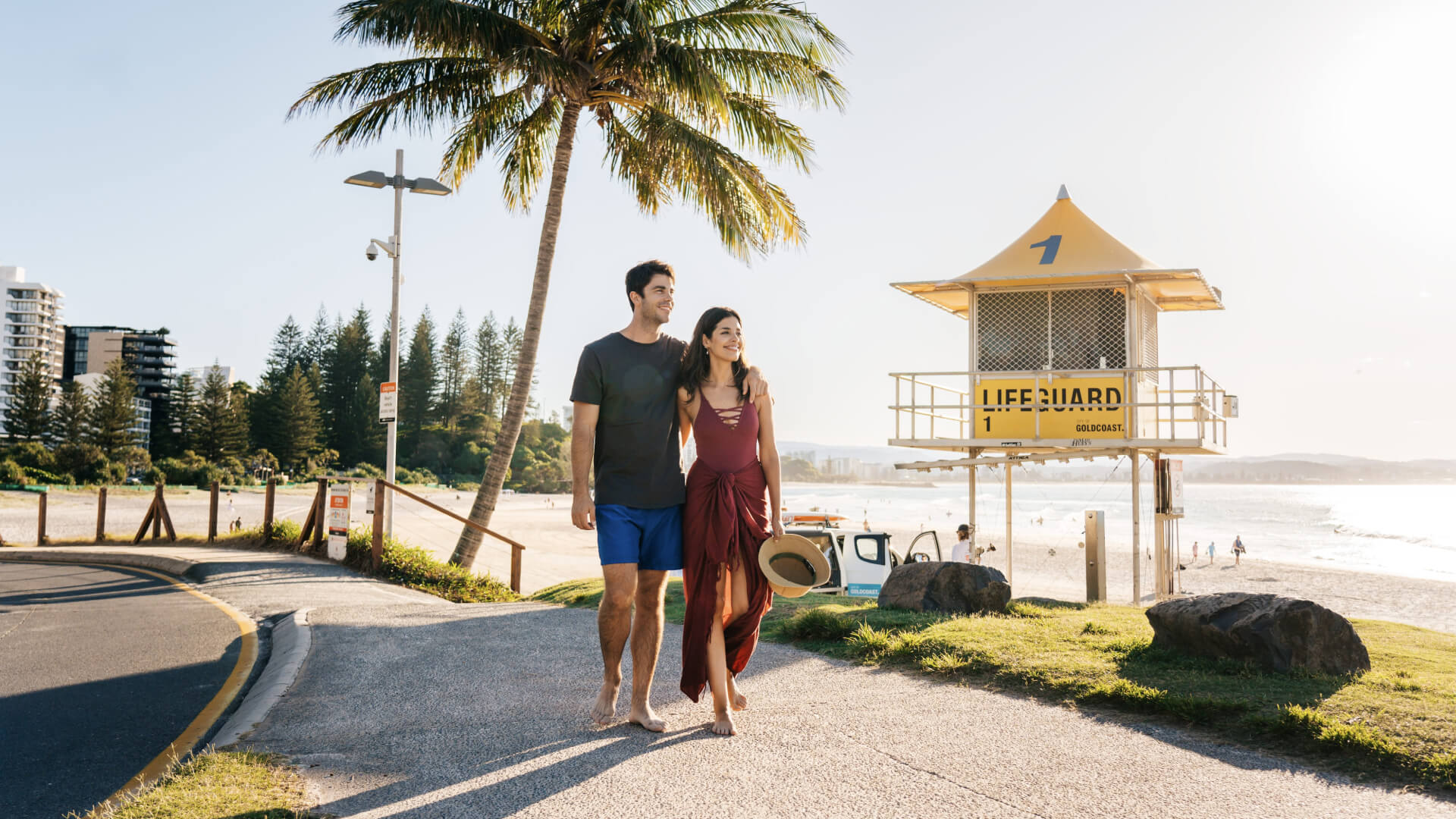 Couple walking along Coolangatta Beach at sunset past a yellow lifeguard tower with palm trees and ocean views in the background.