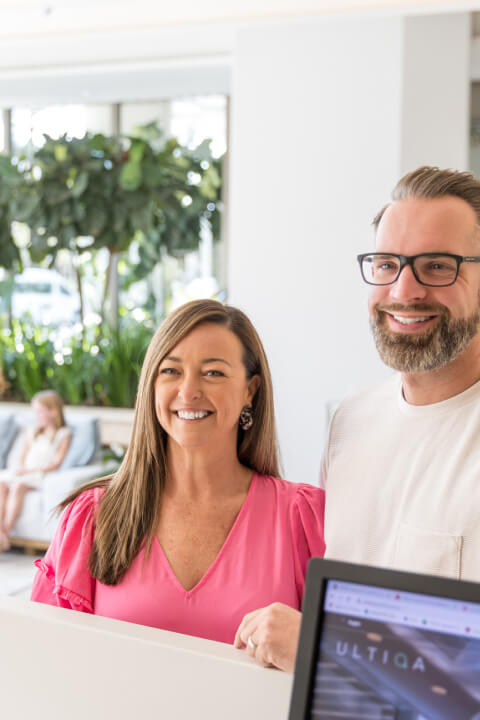 Couple smiling while checking in at a hotel reception, greeted by a friendly staff member.