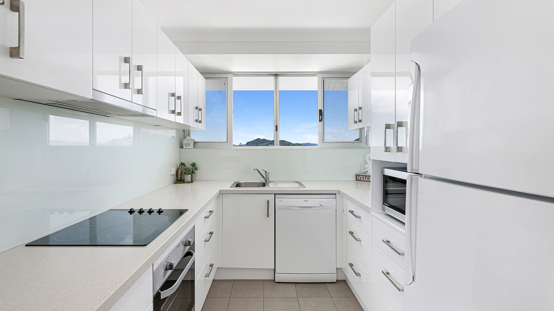 Modern kitchen in a 2 Bedroom Superior Ocean View Apartment featuring white cabinetry, stainless-steel appliances & window with ocean & hillside views.