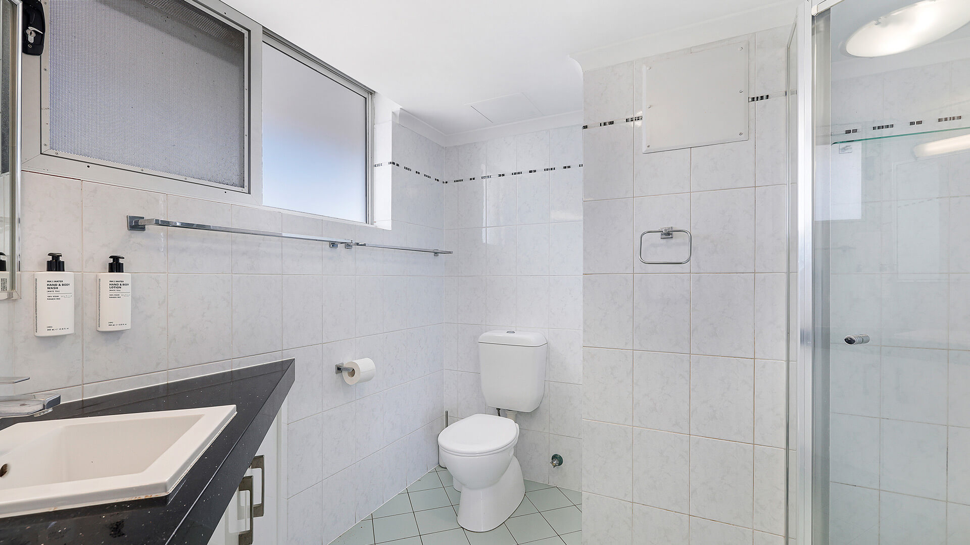 Bathroom in the 2 Bedroom Superior Ocean View Apartment with a glass-enclosed shower, white vanity, toilet & frosted windows providing natural light.