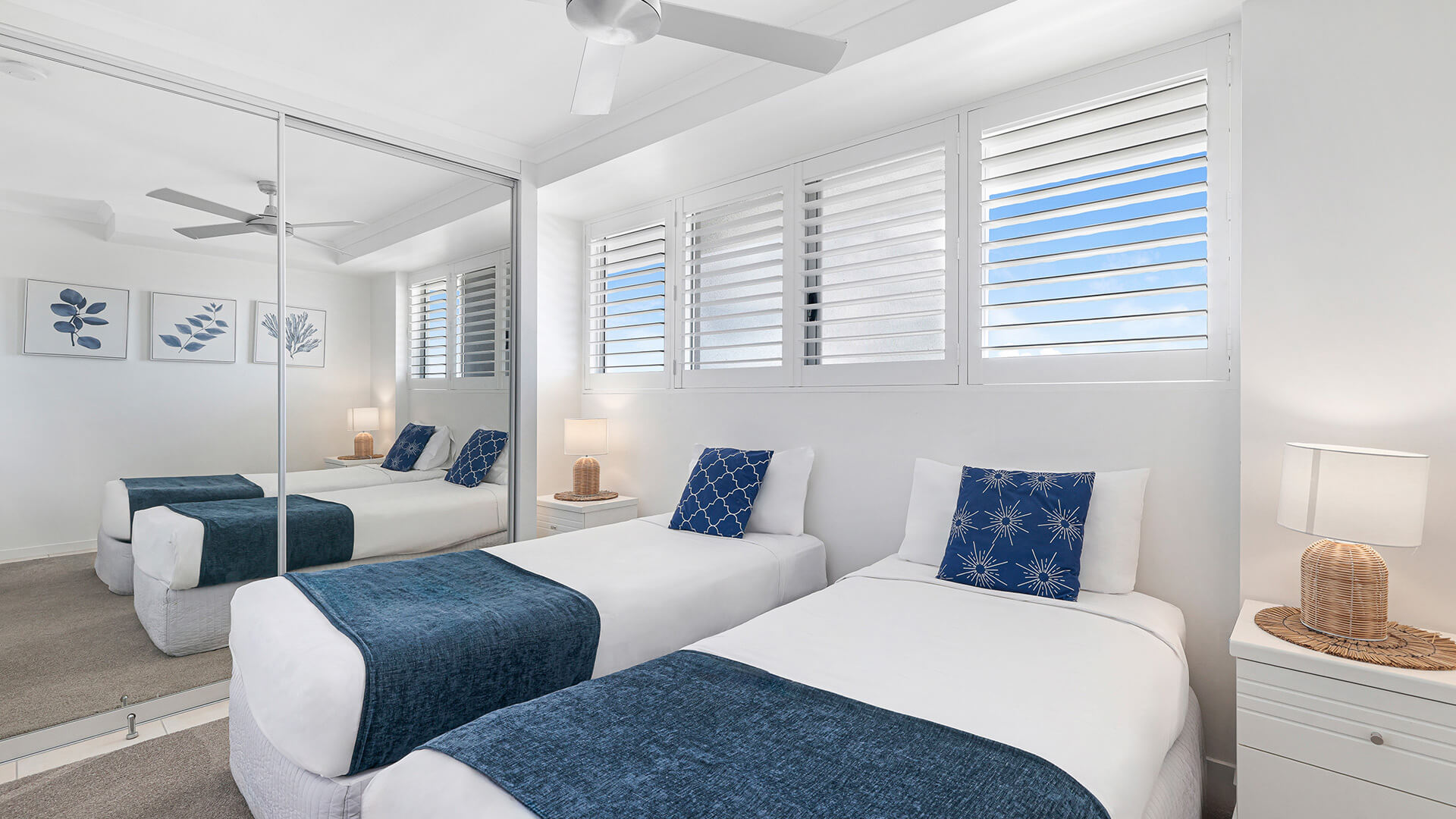 Twin bedroom in a 2 Bedroom Superior Ocean View Apartment with 2 single beds, blue throws, patterned cushions, mirrored wardrobes & shuttered windows.