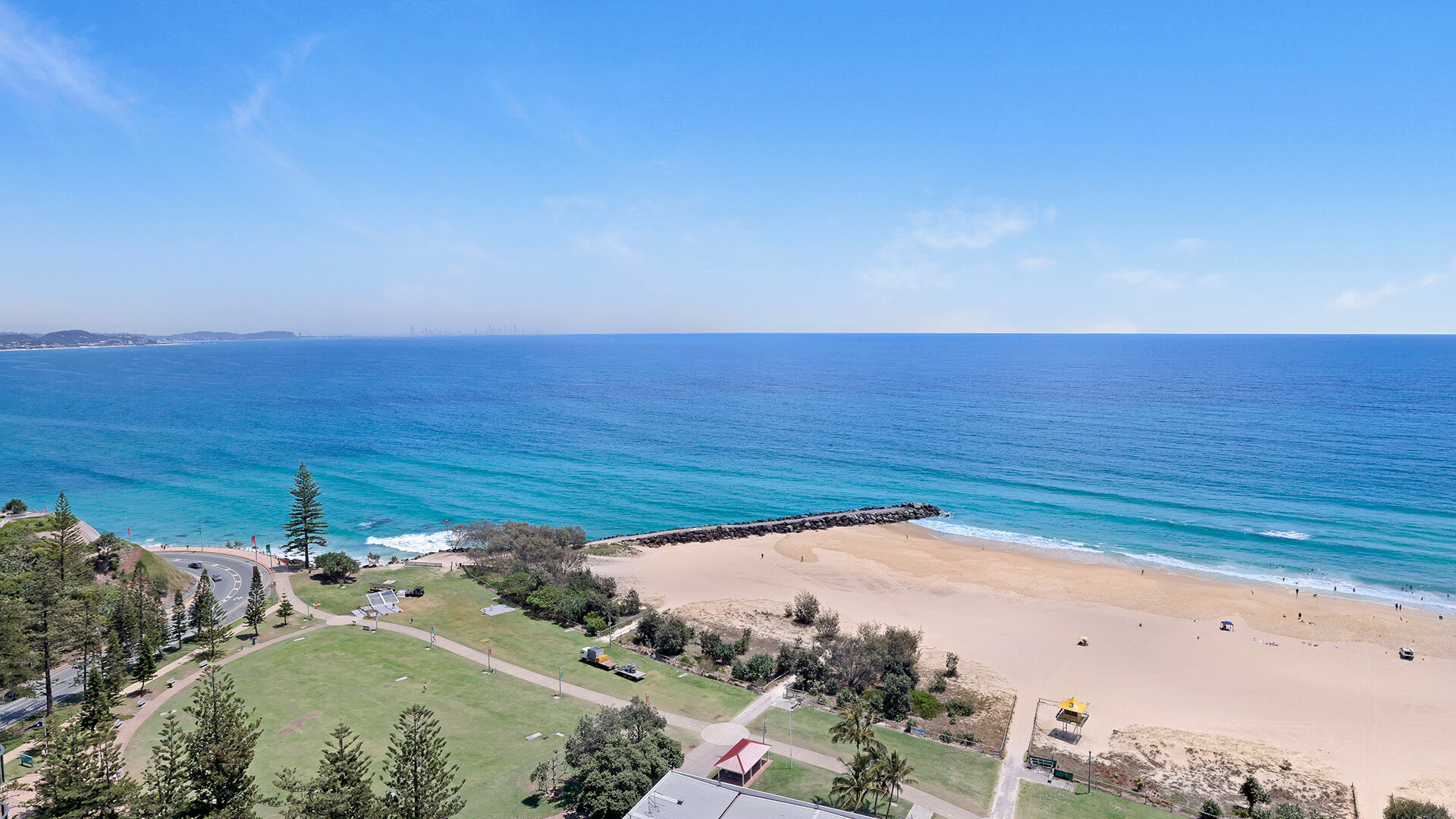Expansive beach and ocean view featuring golden sand, turquoise water, and a grassy park area along the coastline.