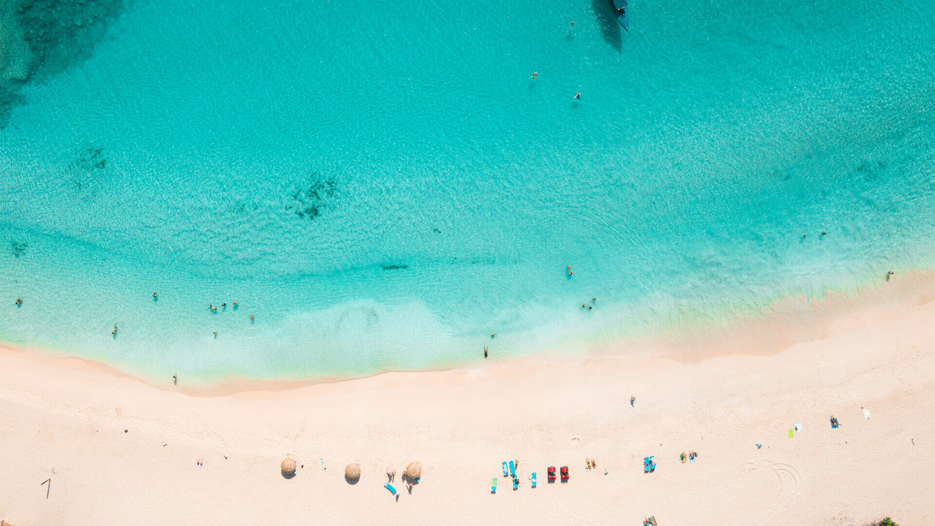 Aerial view of a turquoise ocean meeting a sandy beach with people swimming and relaxing along the shoreline.